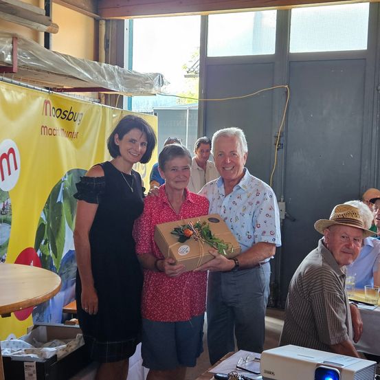 Drei Personen stehen vor einem gelben Banner. Eine Frau in einem schwarzen Kleid und ein Mann in einem Blumenhemd halten eine Geschenkbox. Eine ältere Frau in einem roten Oberteil lächelt. Ein Mann in einem Hut sitzt in der Nähe.