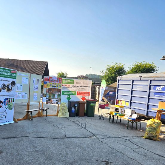 Eine Sammlung von Bannern und Informationstafeln ist im Freien ausgestellt. Es gibt Mülleimer, eine Bank und ein Tisch mit Gegenständen darauf. Ein blauer Container befindet sich auf der rechten Seite. Bäume und ein Haus sind im Hintergrund.