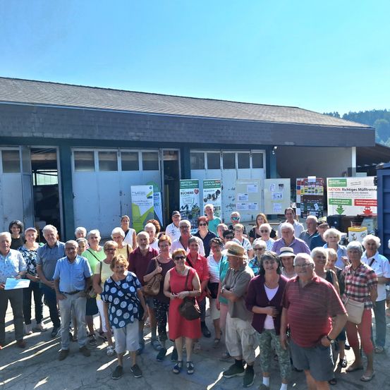 Eine Gruppe von Menschen steht vor einem Gebäude mit Bannern. Sie sind lässig gekleidet, einige tragen Hüte und einige halten Papiere. Das Gebäude hat offene Türen und Fenster. Der Himmel ist klar.