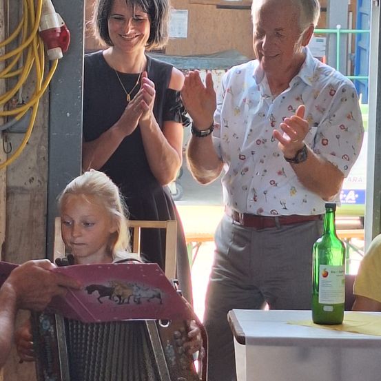 Ein älterer Mann und eine Frau klatschen vor einem kleinen Mädchen, das Akkordeon spielt. Eine grüne Flasche steht auf einem Tisch in der Nähe.