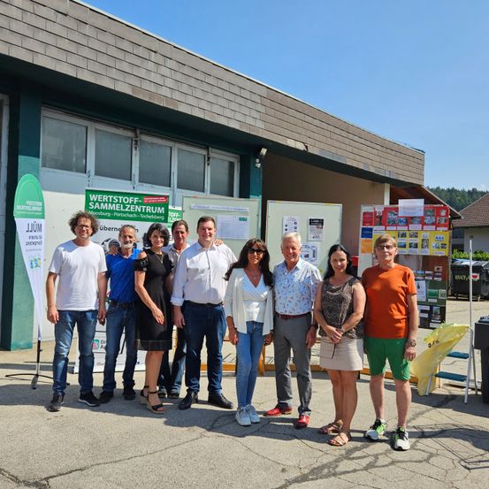 Eine Gruppe von Menschen steht vor einem Gebäude mit der Aufschrift 'Wertstoff-Sammelzentrum Moosburg-Purtschach-Techelsberg'. Sie sind lässig gekleidet und scheinen für ein Foto zu posieren.