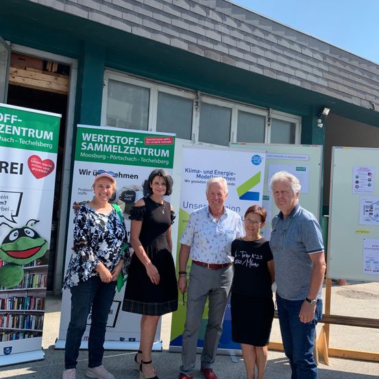 Fünf Personen stehen vor einem Gebäude mit verschiedenen Bannern. Die Frau links trägt ein Blumenoberteil und Jeans. Die zentrale Frau trägt ein schwarzes Kleid. Der zentrale Mann trägt ein Blumenhemd, und die beiden Männer rechts tragen Freizeithemden.