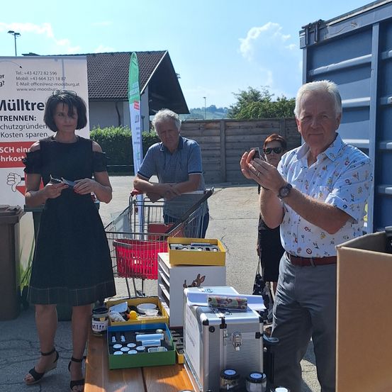 Drei Personen stehen vor einem Tisch mit mehreren Gegenständen, einer hält ein Telefon, ein anderer ein kleines Gefäß. Dahinter steht ein Gebäude mit einem Schild.