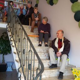 Eine Gruppe von Menschen, darunter ein Mann und mehrere Frauen, sitzen auf den Treppen eines Hauses. Einige lächeln und eine Frau hält einen Becher. Die Treppe hat ein Metallgeländer.