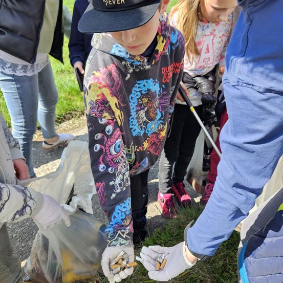 Eine Gruppe von Menschen steht in einem Grasbereich. Sie sammeln Müll auf, tragen Handschuhe und halten Plastiktüten. Die Person vorne trägt eine Mütze und ein farbenfrohes Langarmshirt.