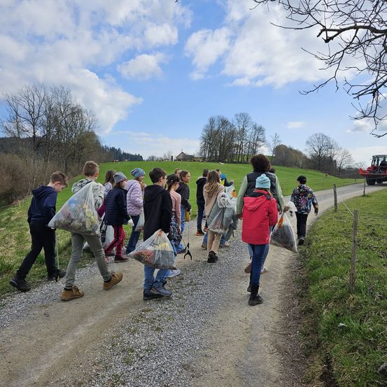 Eine Gruppe von Menschen, die einen Schotterweg hinuntergehen und Taschen tragen. Das Gebiet ist von Grün umgeben. In der Ferne befindet sich ein Traktor auf einem Grasfeld.