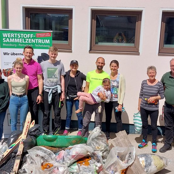 Eine Gruppe von Menschen posiert für ein Foto vor einem Gebäude mit einem Schild, das 'Wertstoff-Sammelzentrum' steht. Sie stehen neben Säcken voller Abfall.