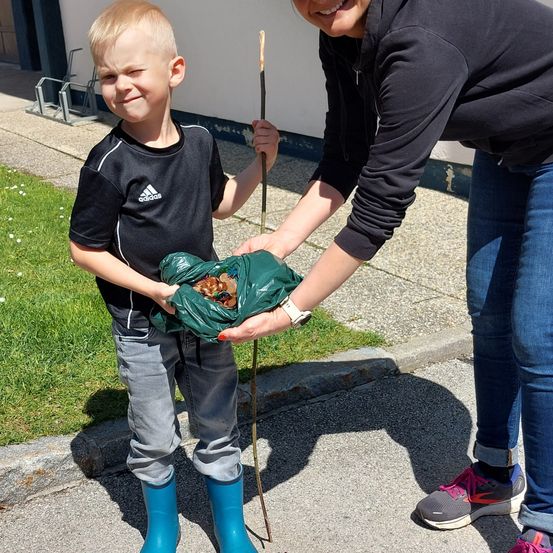 Ein junger Junge mit blauen Stiefeln hält eine grüne Tasche voller Münzen, während eine Frau ihm hilft. Sie stehen auf einem Betonsteig neben einem Rasenbereich.