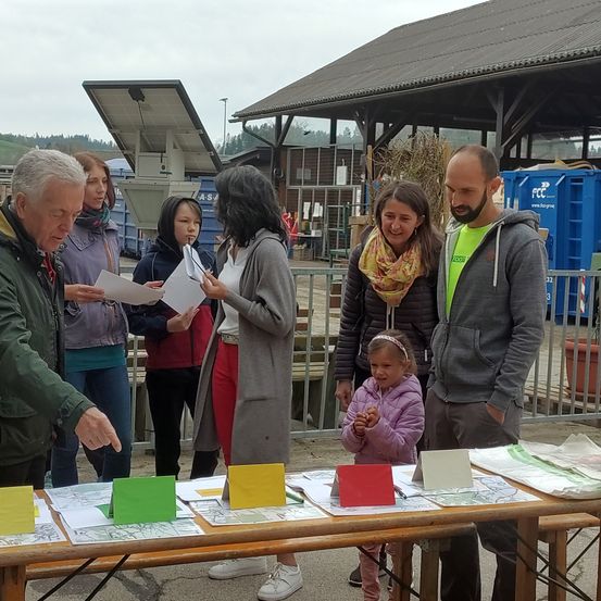 Eine Gruppe von Menschen, darunter Erwachsene und Kinder, steht um einen Tisch mit Papieren und Karten. Eine Frau hält einen Stift und ein Mann zeigt auf den Tisch. Ein Gebäude mit Solarpanelen steht dahinter.