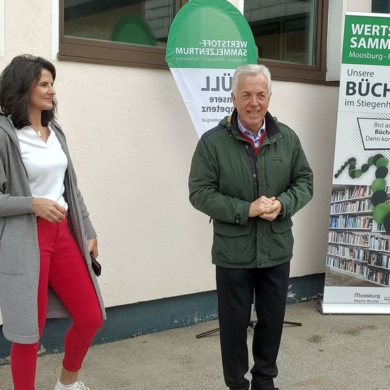 Zwei Menschen stehen vor einem Gebäude. Eine Frau trägt eine graue Jacke und rote Hosen. Ein Mann trägt eine grüne Jacke und schwarze Hosen. Dahinter befindet sich ein Banner und ein Plakat.