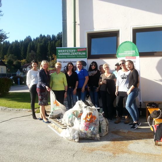 Eine Gruppe von Menschen, einige mit Taschen, steht vor einem Gebäude mit einem Schild, das 'Wertstoff Sammelzentrum' besagt. Sie nehmen an einer Recycling-Veranstaltung teil.