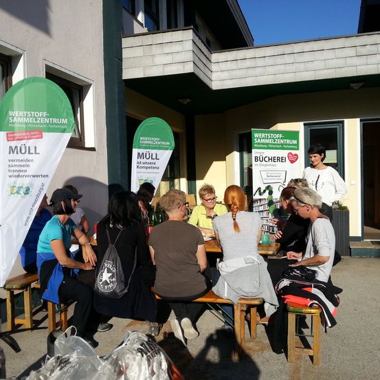 Eine Gruppe von Menschen sitzt auf Bänken vor einem Gebäude und ist in einer Aktivität vertieft. Banner und Plakate sind angebracht, und eine Frau steht in der Nähe. Das Gebäude hat eine Glastür und Fenster.