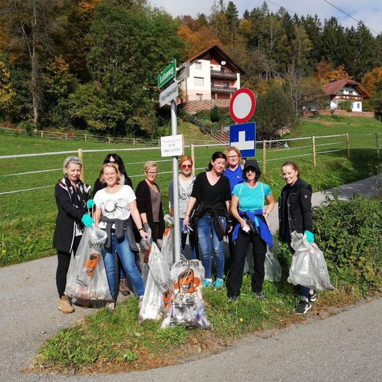 Eine Gruppe von Frauen steht am Straßenrand, trägt Handschuhe und hat Plastiktüten bei sich. Sie posieren für ein Foto, wahrscheinlich nach einer Aufräumaktion. Im Hintergrund befinden sich Häuser, Bäume und ein Zaun.