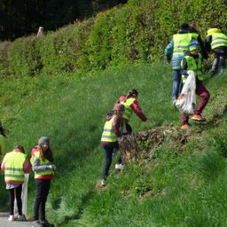 Eine Gruppe von Freiwilligen in reflektierenden Westen und Handschuhen räumt einen Bereich auf einem Grashang auf. Einige sammeln Müll, während andere gehen. Der Himmel ist klar.