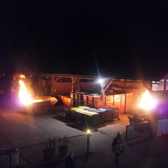 In der Nacht brennt ein Feuer in einem Müllcontainer eines Lagerhauses. Zwei Personen sind anwesend, eine trägt die Uniform eines Feuerwehrmanns.