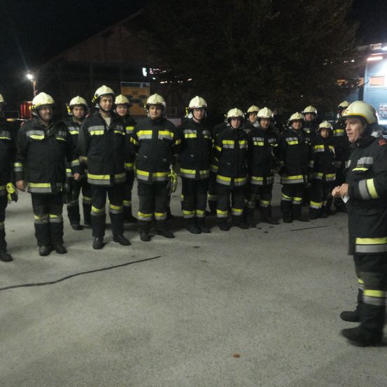 Eine Gruppe von Feuerwehrleuten in voller Uniform steht nachts in einer Reihe, wobei eine Person sie anspricht.