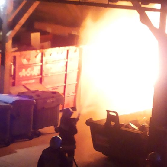 Feuerwehrleute bekämpfen einen Brand in einem Lagerhaus. Rauch und Flammen sind sichtbar, daneben befinden sich Müllcontainer.