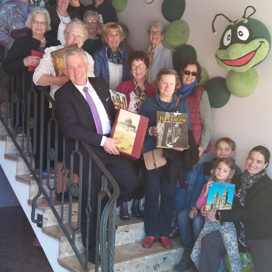 Eine Gruppe von Menschen, einschließlich Erwachsener und Kindern, steht auf einer Treppe. Sie halten Bücher und lächeln für ein Foto. Der Mann in der Mitte hält ein Buch mit dem Titel 'Jerusalem.' Hinter ihnen ist eine Wand mit grünen und weißen Dekorationen.