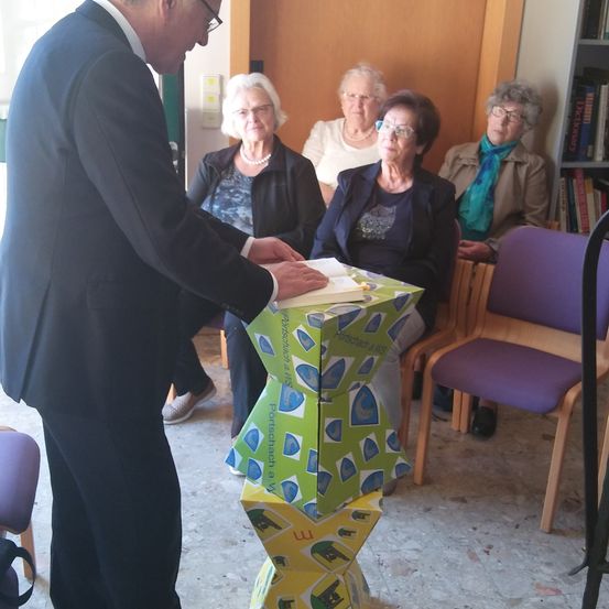 Ein Mann in einem Anzug überreicht einer Frau, die zwischen anderen älteren Frauen sitzt, eine Geschenkbox in einem Raum mit Bücherregalen und Stühlen.