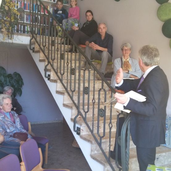 Eine Gruppe von Menschen sitzt auf einer Treppe und hört einem Mann im Anzug zu. Sie sind in einem Raum mit Bücherregalen und grünen Dekorationen.