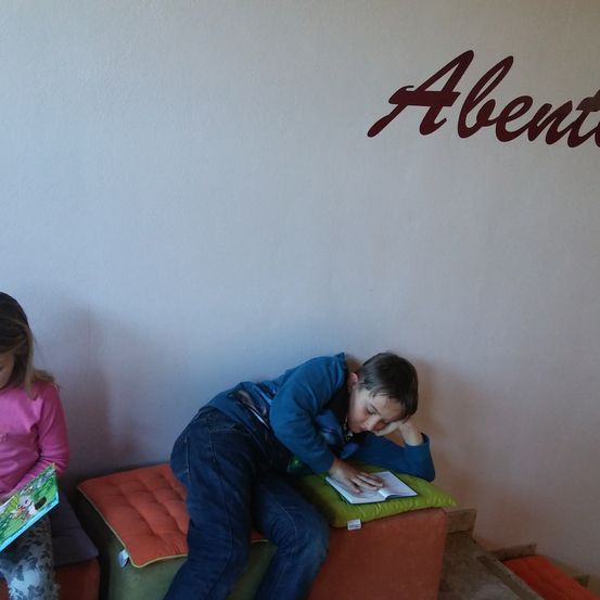 Zwei Kinder sind in einem Raum. Ein Mädchen sitzt auf einem Kissen und liest ein Buch. Ein Junge schläft auf einem anderen Kissen mit einem Buch im Schoß. An der Wand hinter ihnen steht ein rotes Wort 'Abentu'.