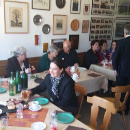 Eine Gruppe von Menschen sitzt in einem Restaurant an einem Tisch und isst und trinkt. Es gibt mehrere Teller, Tassen und Flaschen auf dem Tisch. Die Wände sind mit verschiedenen Rahmen und Bildern dekoriert.