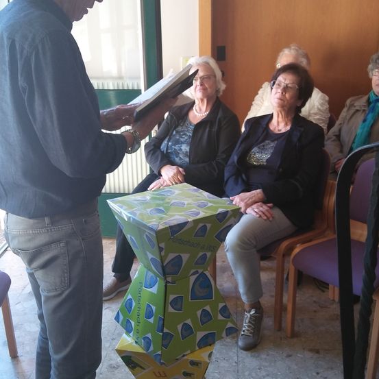 Ein Mann liest ein Buch vor einer Gruppe älterer Frauen, die auf Stühlen sitzen, eine Frau trägt eine Halskette. Ein verpacktes Geschenk liegt vor ihnen.