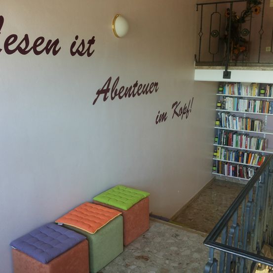 Eine weiße Wand mit dem Text 'Lesen ist Abenteuer im Kopf!' neben drei gepolsterten Hockern. In der oberen rechten Ecke befindet sich ein schwarzer Handlauf mit einem Blumenstrauß. Rechts ist ein Holzregal mit Büchern gefüllt.