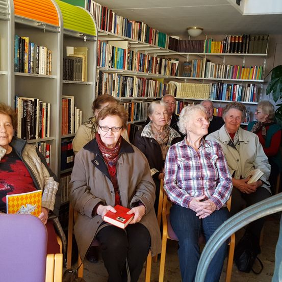 Mehrere ältere Frauen sitzen in Stühlen in einer Bibliothek und schauen nach vorne. Sie halten Bücher und tragen Brillen und Schals. Die Bibliothek hat weiße Regale, die mit Büchern gefüllt sind.