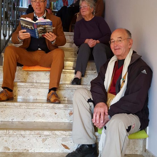 Drei ältere Personen sitzen auf der Treppe. Ein Mann liest ein Buch, während eine Frau neben ihm sitzt. Ein weiterer Mann sitzt rechts und trägt eine Brille und einen Schal.