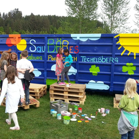 Eine Gruppe von Kindern steht auf Holzpaletten neben einem blauen Container mit der Aufschrift 'Sau BÃ¤RES'. Einige von ihnen malen, während andere herumstehen.