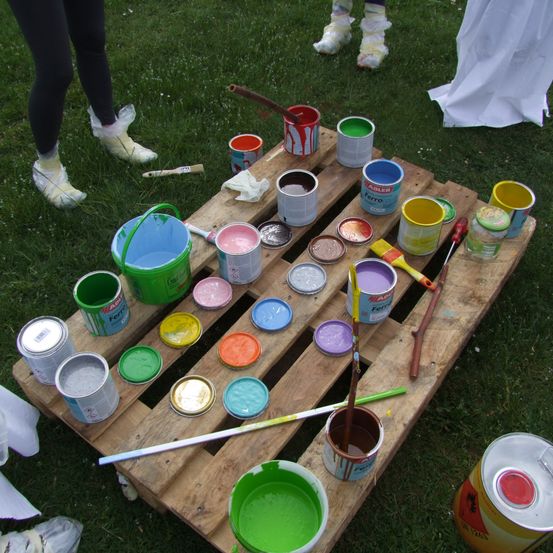 Ein Holzpalette mit verschiedenen Farbdosen und Eimern, Pinseln und einem offenen grünen Eimer auf dem Rasen.