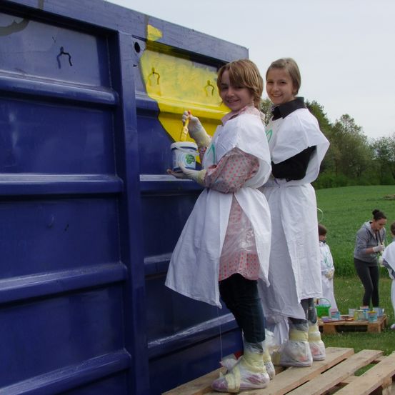 Zwei Mädchen in Schutzkleidung malen einen blauen Müllcontainer gelb mit einem Pinsel. Andere Leute malen um sie herum.
