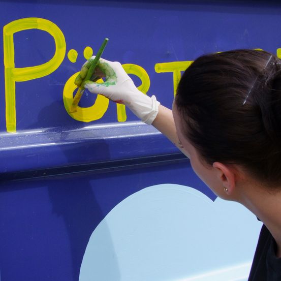Eine Frau in einem weißen Handschuh malt einen gelben Buchstaben P auf eine blaue Oberfläche. Sie hält einen Pinsel und konzentriert sich auf ihre Aufgabe.