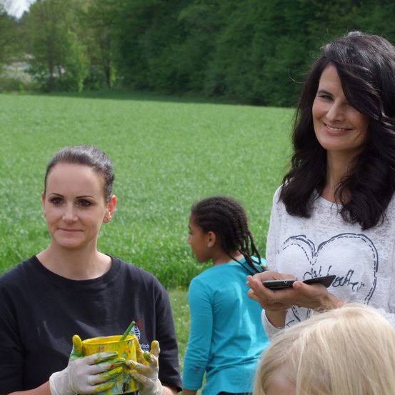Zwei Frauen stehen auf einem Feld, eine hält einen gelben Farbkübel. Ein Mädchen mit geflochtenen Haaren steht hinter ihnen, alle lächeln. Im Hintergrund benutzt eine Frau ihr Handy.