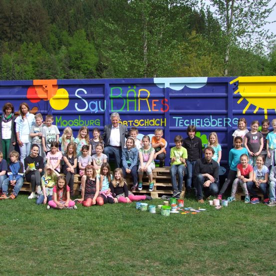 Gruppenfoto von Erwachsenen und Kindern vor einem bemalten Container. Sie sitzen auf Holzpaletten, einige halten Farbdosen. Hinter ihnen ist eine farbenfrohe Wandmalerei.