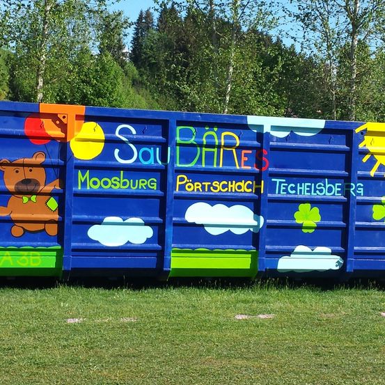 Ein blauer Container ist mit Cartoon-Illustrationen bemalt, darunter ein Bär mit Luftballons und eine Sonne. Der Container trägt den Text 'Saubares Moosburg Portschach Techelsberg'. Er steht auf einem Rasen mit Bäumen im Hintergrund.