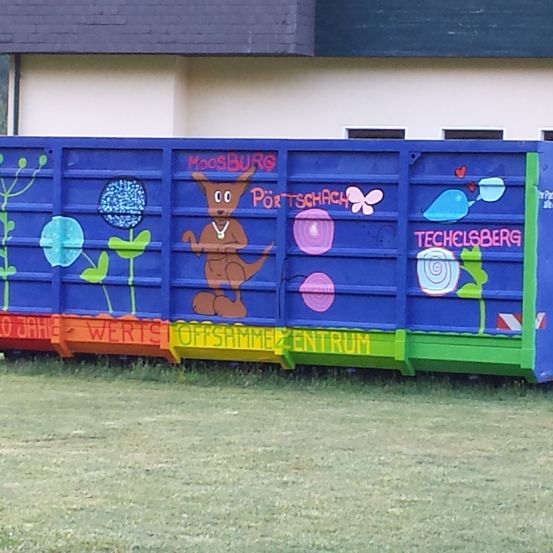 Ein blauer Müllcontainer mit Graffiti steht vor einem Gebäude. Er zeigt einen Kängurus mit Halskette, Blumen, Schmetterlingen und verschiedenen anderen Designs. Der Container trägt auch Text wie 'Moosburg', 'Portschach' und 'Techelsberg'.