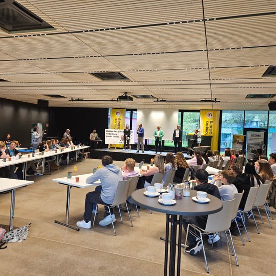 Eine Gruppe von Menschen sitzt in einem Raum mit einer Bühne, einem Banner und einer Leinwand. Die Menschen sitzen in Reihen, der Bühne zugewandt. Die Bühne hat ein Podium und ein Banner. Der Raum hat eine Leinwand, ein Banner und eine Decke mit Lichtern. Die Menschen tragen Freizeitkleidung.