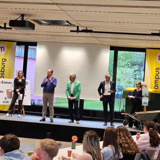 Vier Personen stehen auf einer Bühne mit einem Hintergrund, auf dem 'campus' steht. Eine Frau hält ein Schild. Das Publikum, bestehend aus jungen Leuten, sitzt vor ihnen. Es gibt Blumen in Vasen und Tassen auf einem Tisch vor dem Publikum.