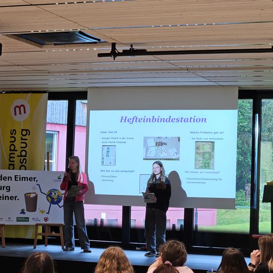 Zwei Frauen stehen auf der Bühne, halten Bücher in den Händen und sprechen in Mikrofone. Eine Projektionsleinwand zeigt den Titel 'Heftbindungsdestation'. Ein Publikum sitzt vor ihnen.