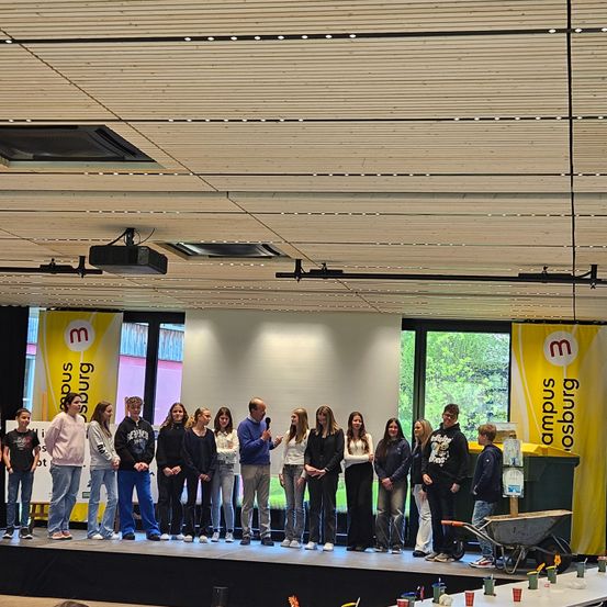 Eine Gruppe von Schülern steht auf einer Bühne vor einem gelben Banner. Einige halten ein Mikrofon, und eine Schubkarre steht in der Nähe. Sie sind lässig gekleidet und scheinen eine Aufführung oder Präsentation abzuhalten.