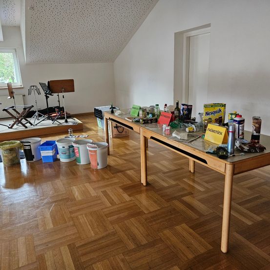 Ein Dachbodenzimmer mit Holzboden. Verschiedene Gegenstände stehen auf den Tischen, darunter eine gelbe Schachtel mit der Aufschrift Nesquick. Es gibt Eimer, ein Stativ und andere Gegenstände auf dem Boden. Ein Heizkörper und ein Fenster befinden sich im Raum.
