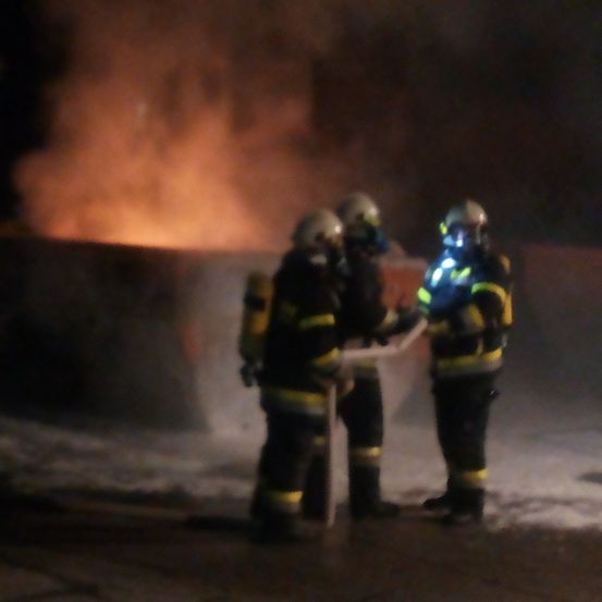 Drei Feuerwehrleute in Schutzkleidung stehen vor einem brennenden Gebäude, einer hält ein Werkzeug, ein anderer wird von einer Taschenlampe beleuchtet.