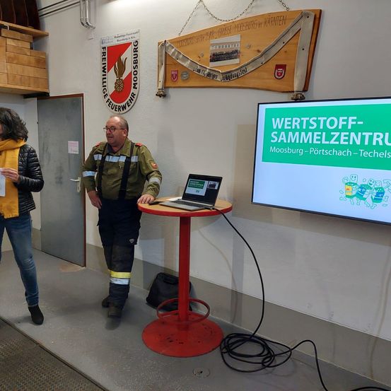 Ein Feuerwehrmann in Uniform steht neben einem Tisch mit Laptop, während eine Frau ein Dokument hält. An der Wand hängen ein Banner und ein Schild mit einem großen Buchstaben 'B'.