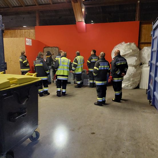 Eine Gruppe von Feuerwehrleuten steht in einem Raum, trägt ihre Uniformen und einige mit reflektierenden Westen. Hinter ihnen ist eine rote Wand und einige weiße Säcke.