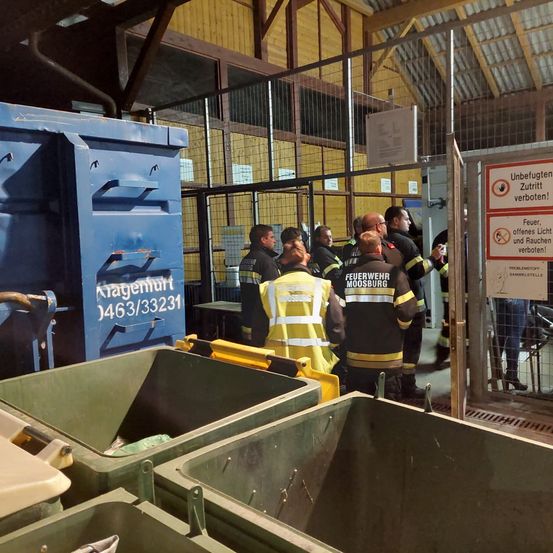 Eine Gruppe von Feuerwehrleuten in Uniform steht in einem Lagerbereich mit großen Behältern und einem Schild, das offenes Feuer und Rauchen verbietet.