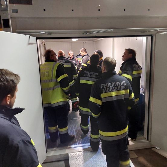 Feuerwehrleute versammeln sich in einem Raum mit einer weißen Tür. Sie tragen gelbe und schwarze Uniformen. Einer von ihnen hält ein rotes Objekt.