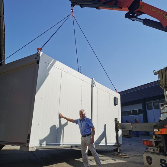 Ein Mann steht neben einer großen weißen Struktur, die von einem Kran unter blauem Himmel gehoben wird.
