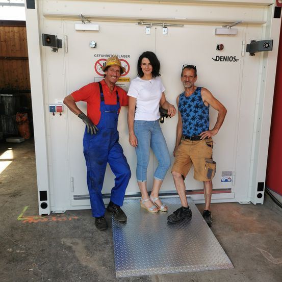 Drei Personen stehen vor einer weißen Tür. Ein Mann trägt einen Hut und Handschuhe, eine Frau ein weißes T-Shirt und ein anderer Mann ein ärmelloses Hemd. Hinter ihnen befindet sich ein Metallboden.